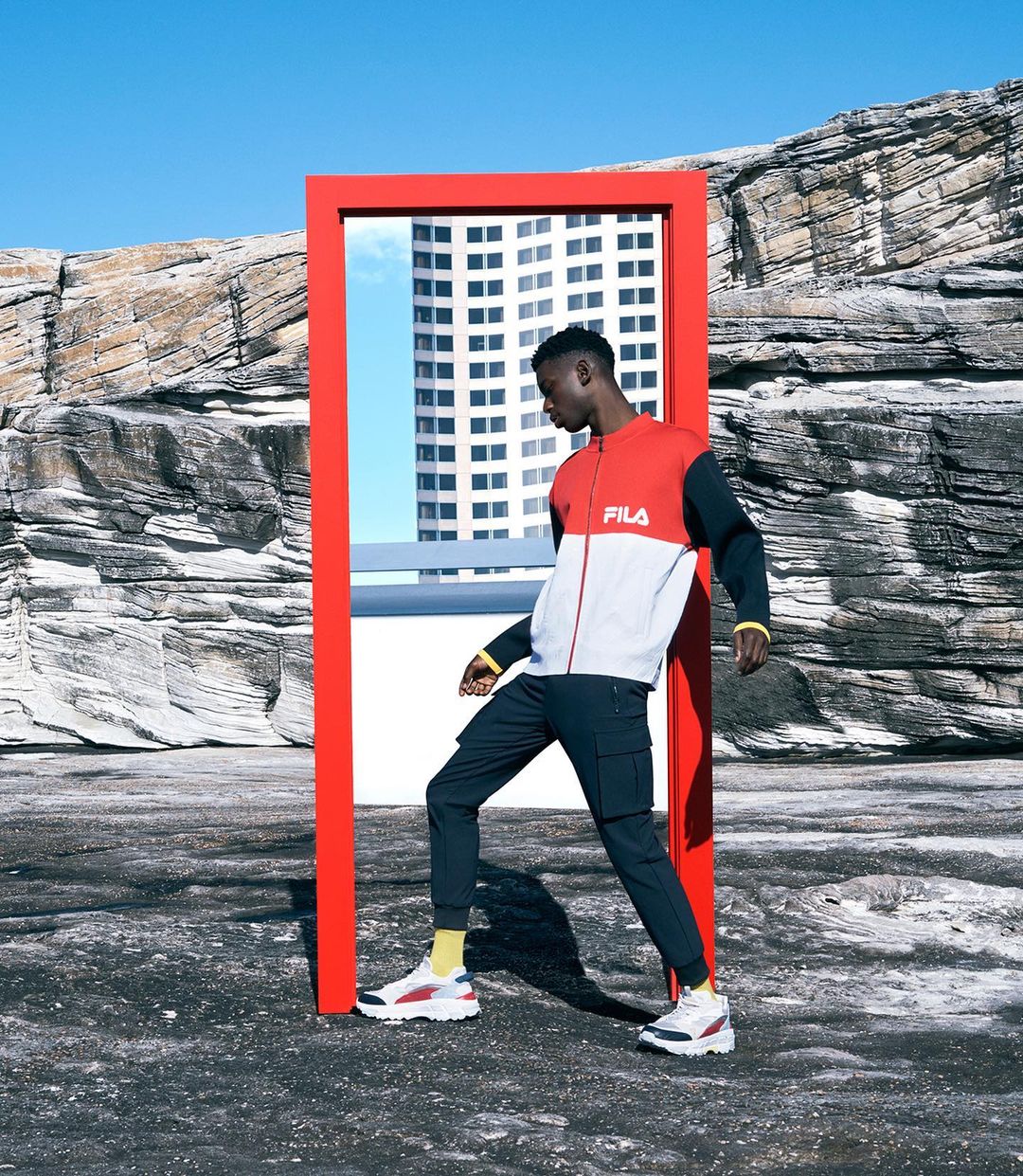 Athletic Black male model showcases sportswear in outdoor setting with minimalist red doorframe, building reflection, and rock formations.