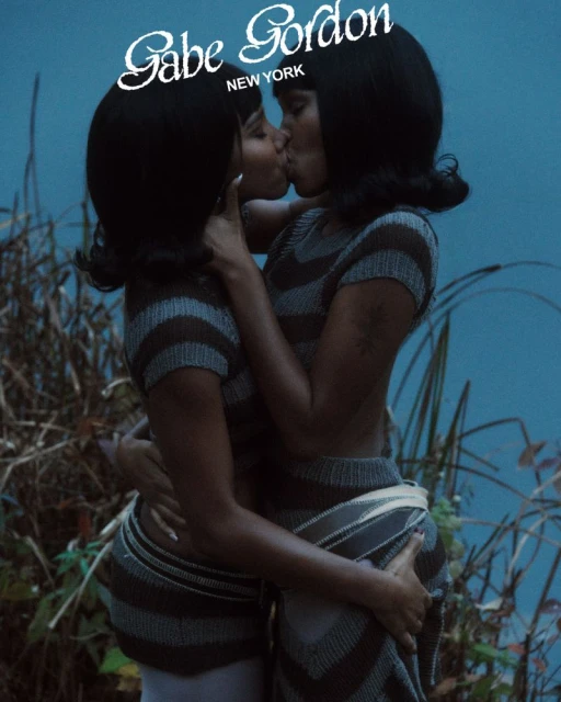 Two adult females kissing in a close-up shot, wearing matching striped knitwear, in front of a blurred background with some vegetation visible.