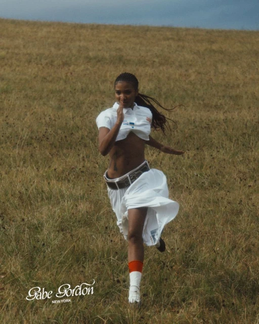 An athletic woman in a white outfit runs across a grassy field under a cloudy sky, showcasing an active outdoor lifestyle and fashion.