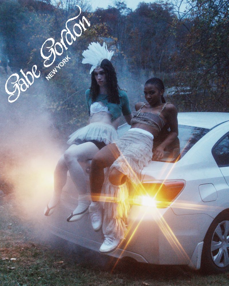 Two women pose on a car in an outdoor setting with a forest backdrop, in a fashion-editorial style with fog and nighttime lighting.