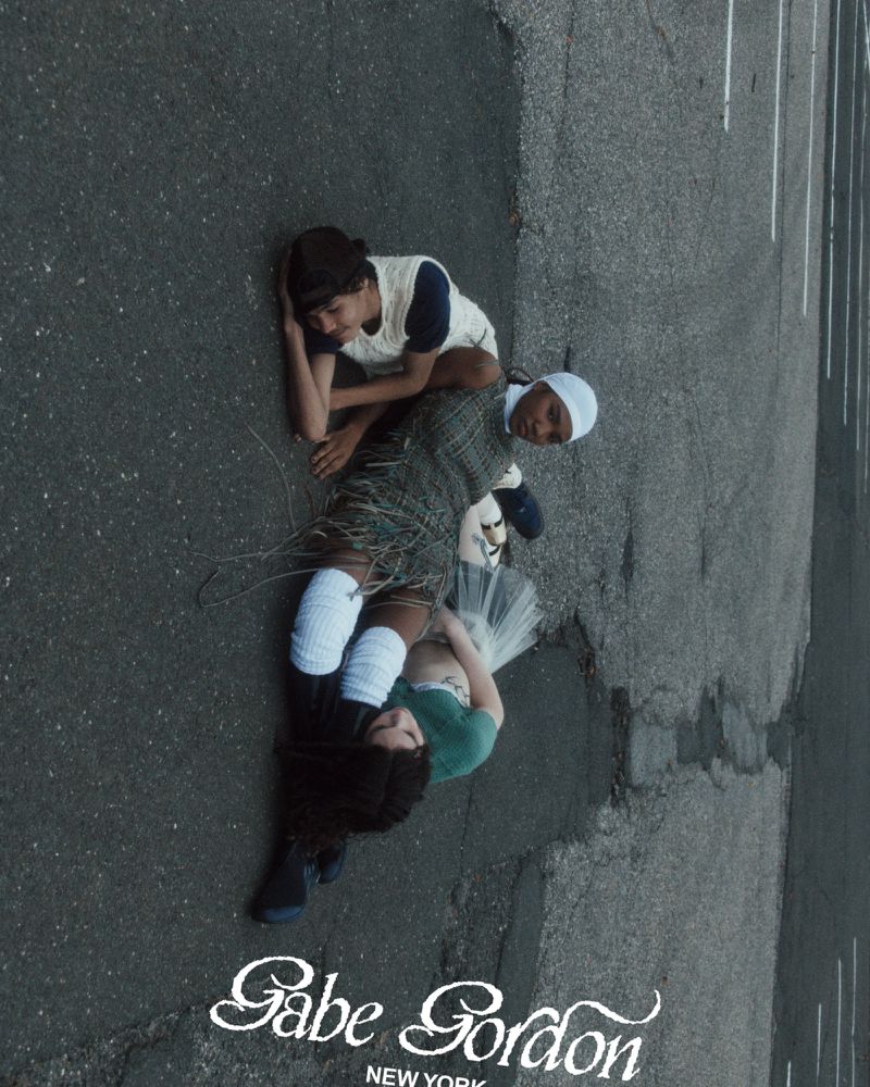 Three young people lie on a paved surface in a group pose. One is wearing a white cap and another, white knee-high socks.