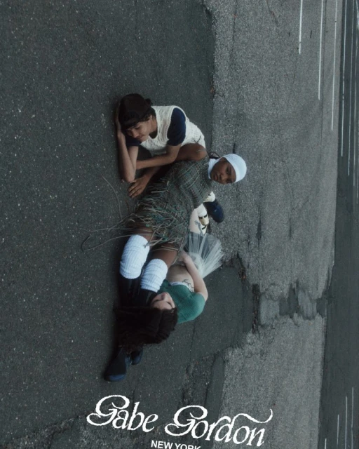 Three young people lie on a paved surface in a group pose. One is wearing a white cap and another, white knee-high socks.