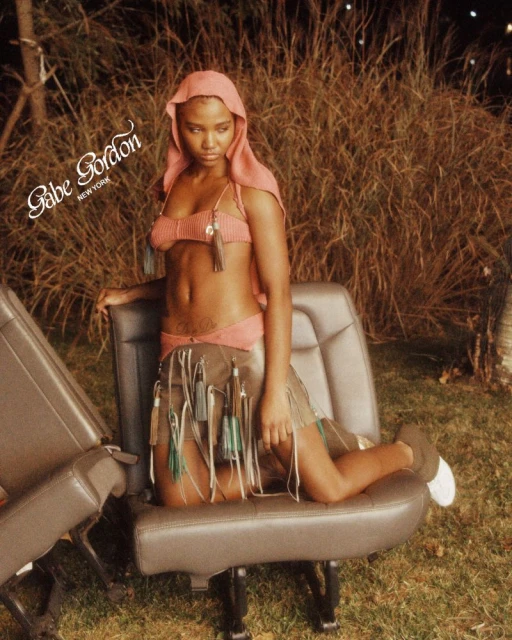 Woman in bikini and skirt poses on car seats in outdoor nighttime portrait. She wears a hood and faces forward with a serious expression.