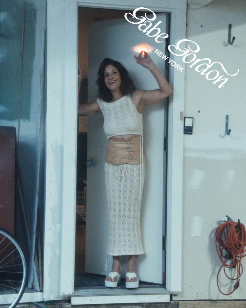 A woman in a knit outfit stands in a doorway with a bicycle wheel, orange cord, and brand logo visible in the background.