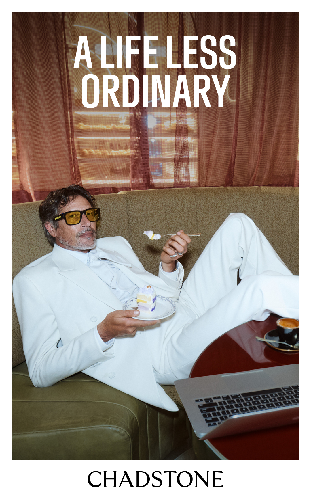 Adult male in a white suit lounges on a green couch, eating cake, with a laptop and coffee table in a casual indoor setting.