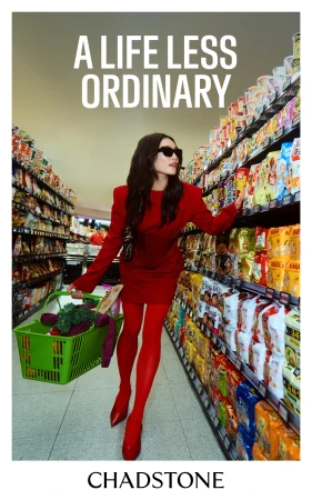 A woman in a red outfit walks through a supermarket aisle, carrying a green shopping basket filled with groceries and snacks.
