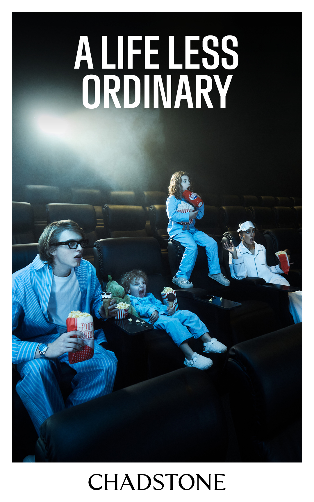 A group of people, including children and adults, sit in an empty cinema wearing pajamas and holding popcorn in a dimly lit setting.