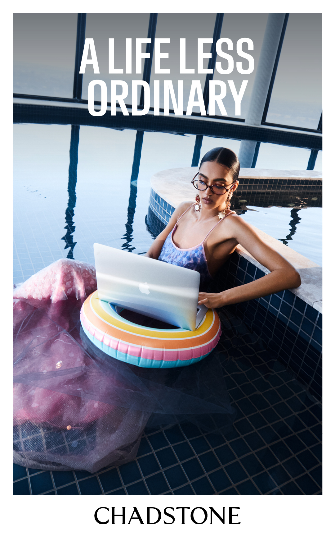 Woman in a pool sits on a floatie using a laptop with the text, "A Life Less Ordinary," visible above her; tile and water textures.