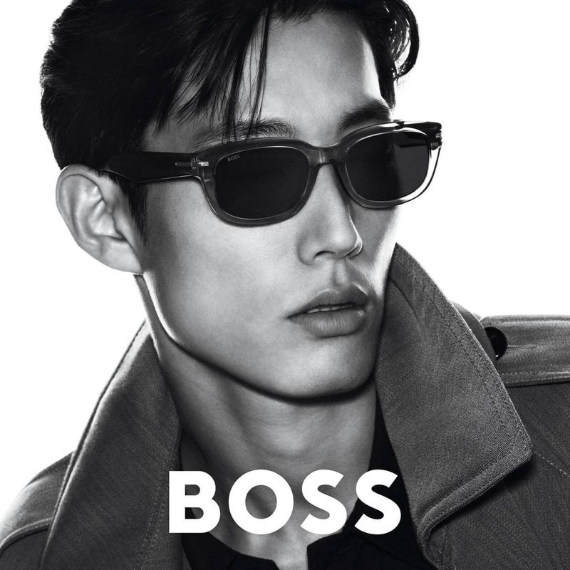 A man poses indoors wearing sunglasses and a collared coat with the brand name BOSS in white text at the bottom of the frame.