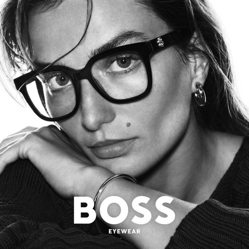 Close-up studio shot of a woman wearing black-framed eyeglasses, a silver bracelet, and hoop earrings, showcasing BOSS Eyewear branding.