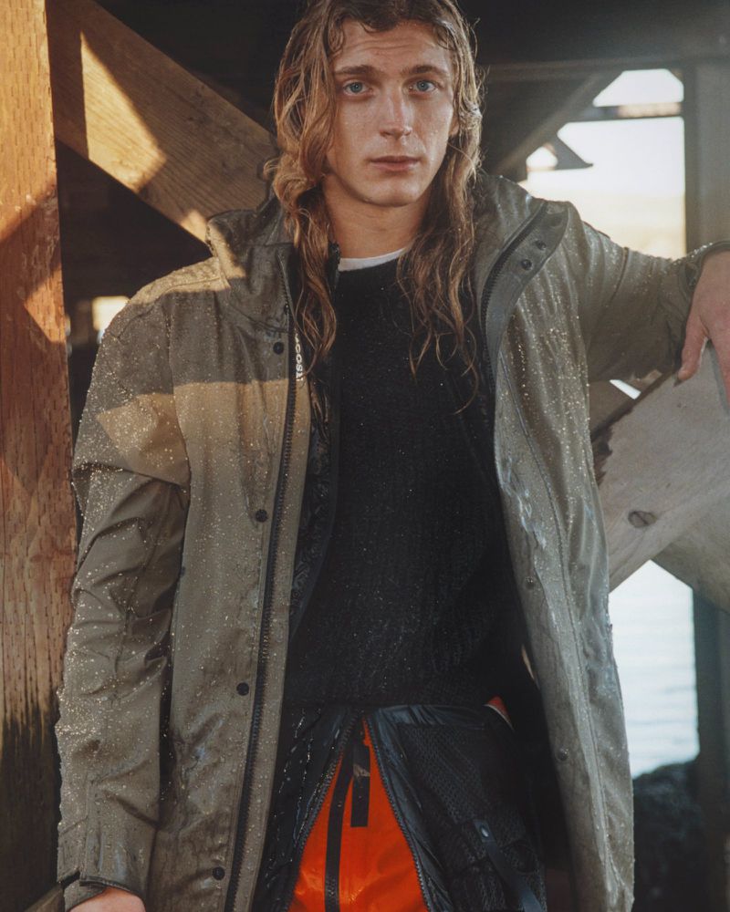 Adult male with long blond hair wears a green hooded raincoat and dark sweater; partially outdoors with wood beams in the background.