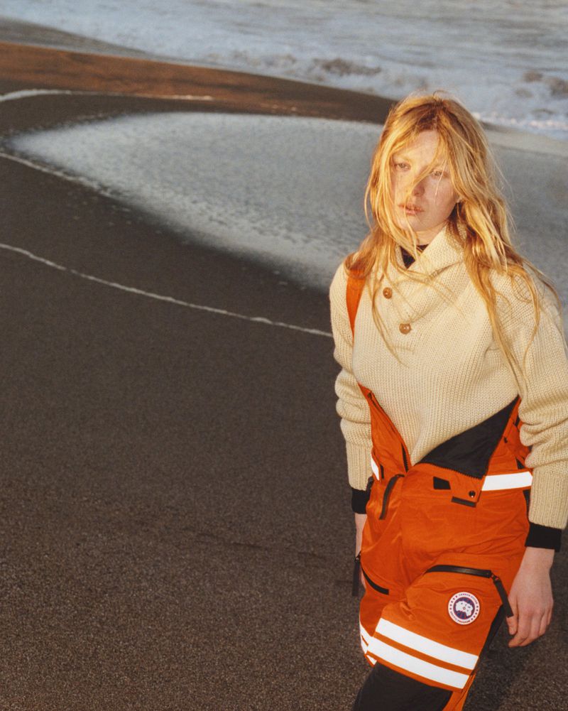 A blonde adult female standing on a beach, wearing an off-white sweater and orange bib overalls with a Canada Goose logo on the left leg.