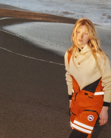 A blonde adult female standing on a beach, wearing an off-white sweater and orange bib overalls with a Canada Goose logo on the left leg.