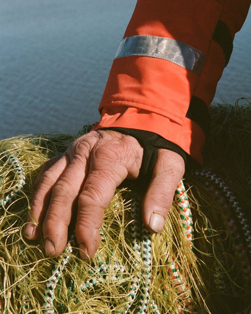 Close-up on an adult male's hand resting on a fishing net, wearing an orange jacket sleeve, against a blurry water background.
