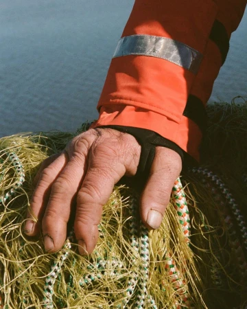 Close-up on an adult male's hand resting on a fishing net, wearing an orange jacket sleeve, against a blurry water background.