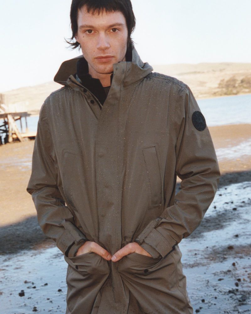 A young adult male stands in a full-length coat on a beach, with a distant body of water and land visible under an overcast sky.