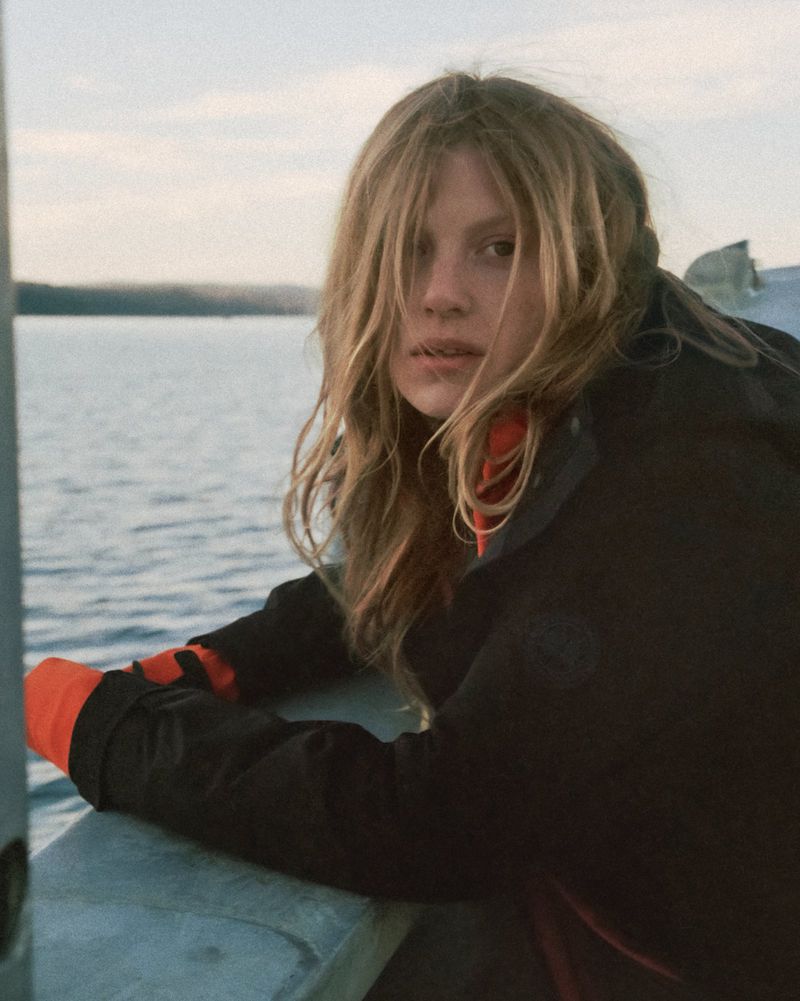 Woman in a black jacket looks at the camera near a lake under a bright sky. She wears an orange undershirt and has blonde hair.