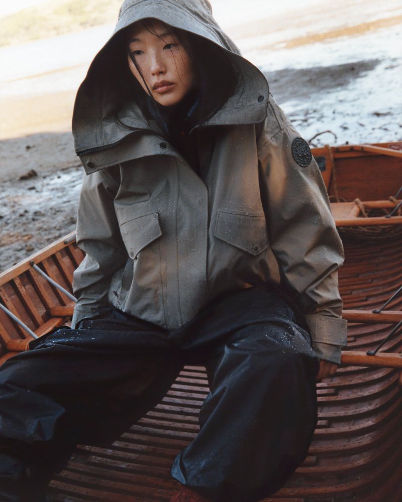 A woman with dark hair in a hooded olive-green jacket sitting in a wooden boat on a beach, looking downwards with a serious facial expression.