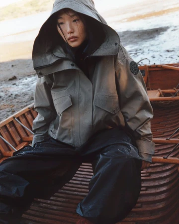 A woman with dark hair in a hooded olive-green jacket sitting in a wooden boat on a beach, looking downwards with a serious facial expression.
