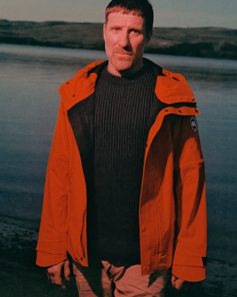Adult male wearing a bright orange hooded jacket and dark sweater posing in front of a lake with rolling hills in the background.