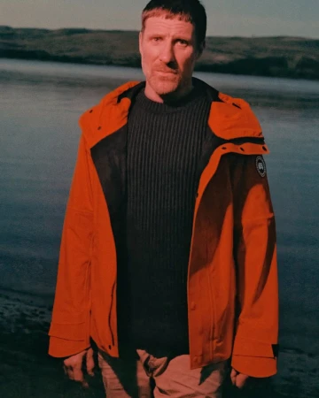 Adult male wearing a bright orange hooded jacket and dark sweater posing in front of a lake with rolling hills in the background.