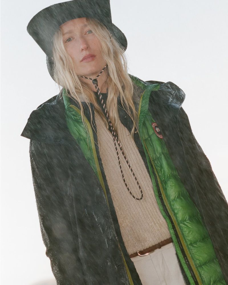 A woman with long blonde hair wearing a bucket hat and outdoor jacket, shot from a low angle with a bright, slightly overexposed background.