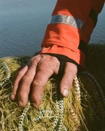 Close-up of a person's hand wearing an orange jacket resting on tangled yellow fishing nets outdoors near water.