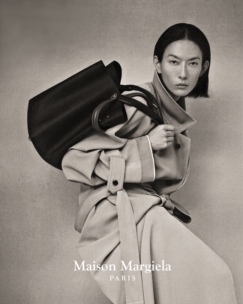 Fashion shot of an Asian woman wearing a beige trench coat with a black handbag over her shoulder, studio shot on gray background.