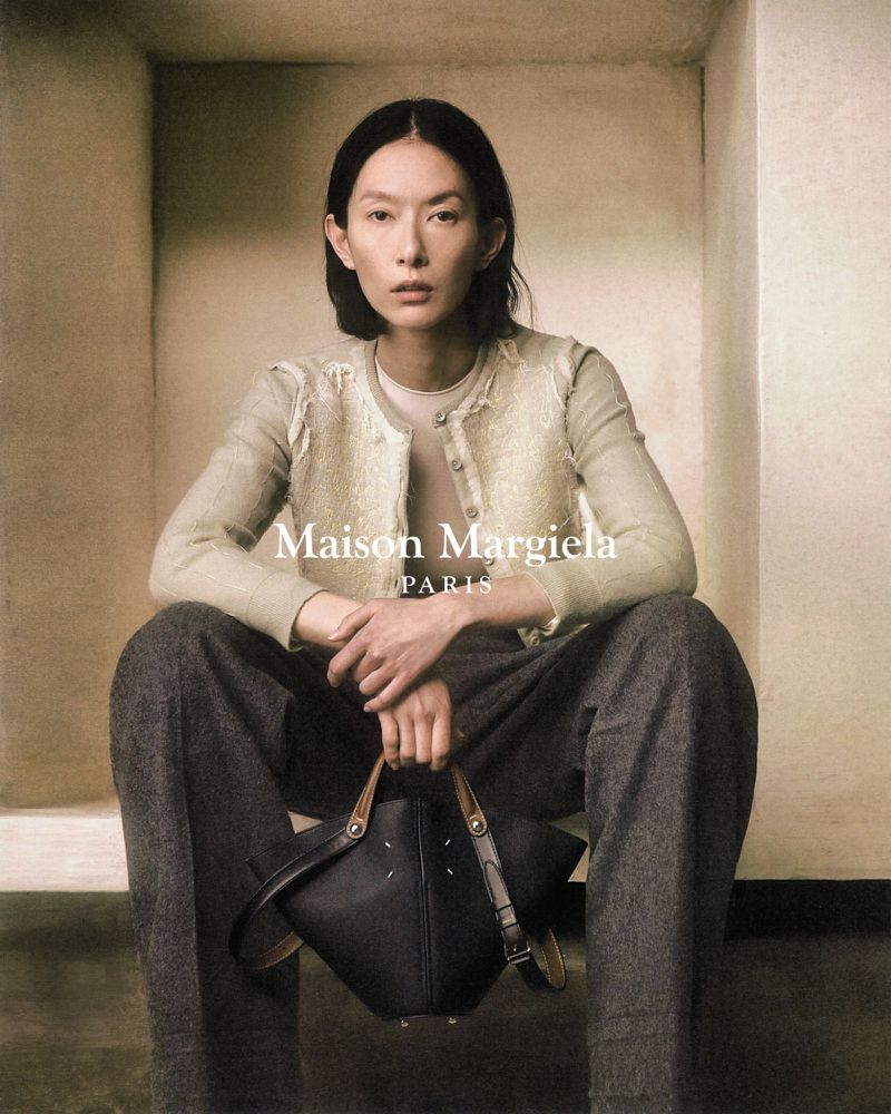 An Asian female model poses indoors with a black leather handbag, wearing gray trousers and a beige cardigan, showcasing high-fashion apparel.