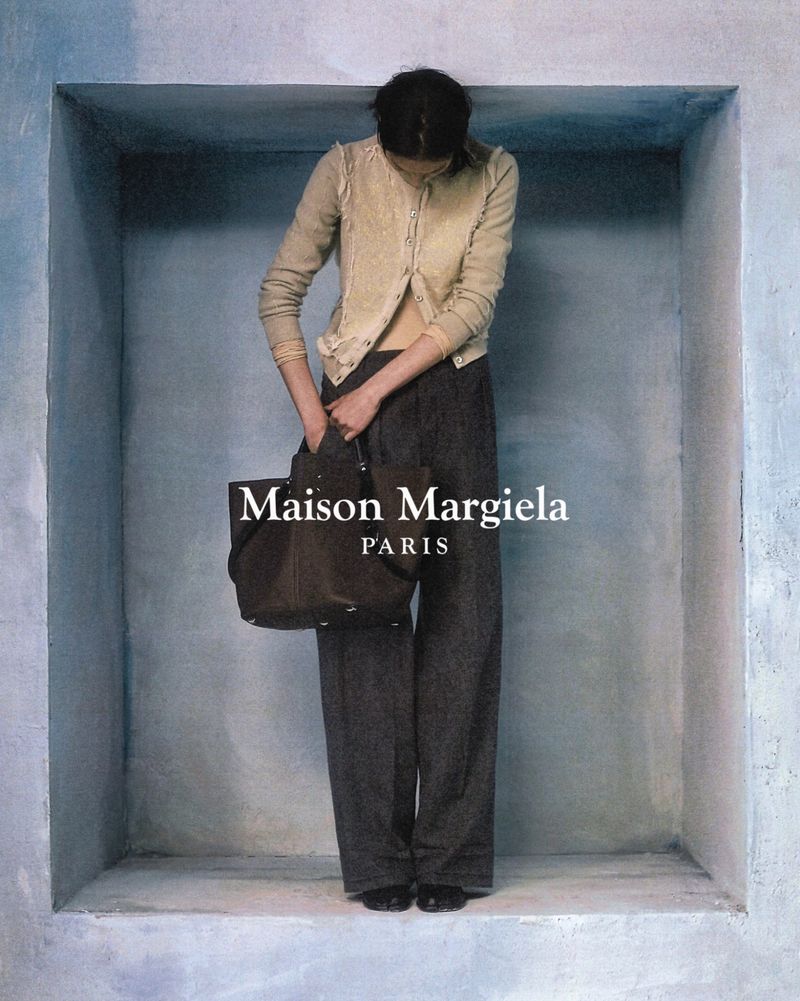 Adult female fashion model stands in a concrete cubicle wearing a tan sweater, grey pants, and holding a black tote bag indoors.