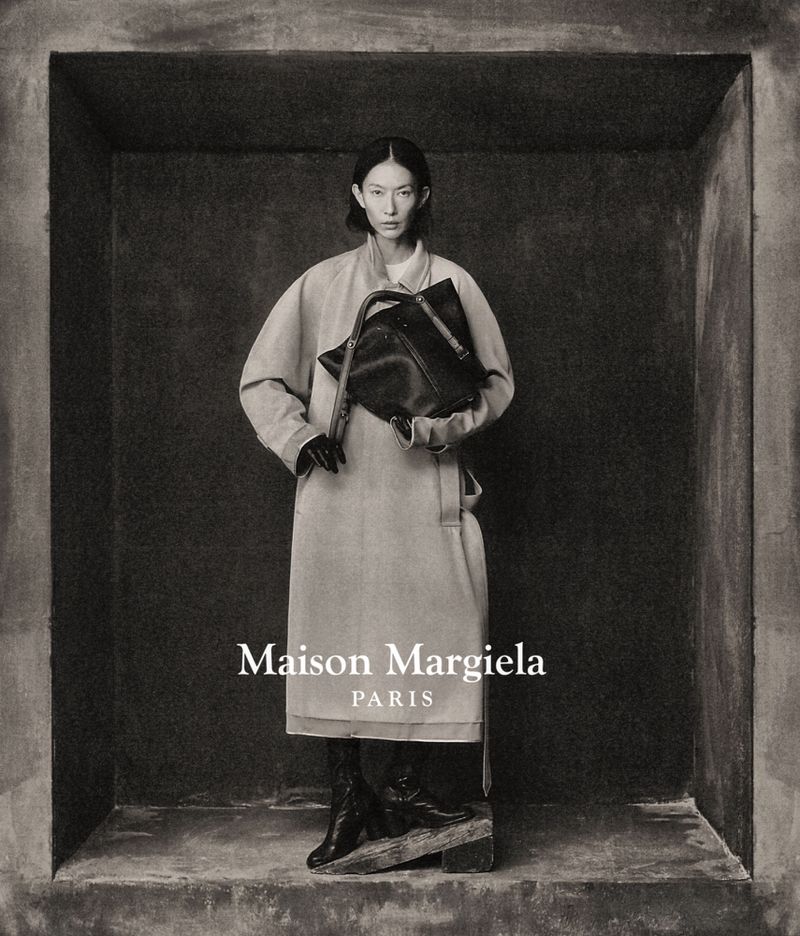 Fashion shot features a woman in a beige coat and black boots, holding a black handbag in a constructed studio setting, monochrome.