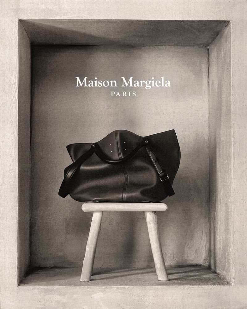 Maison Margiela black leather handbag displayed on a three-legged stool against a textured grey wall in a recessed space.