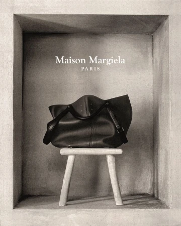 Maison Margiela black leather handbag displayed on a three-legged stool against a textured grey wall in a recessed space.