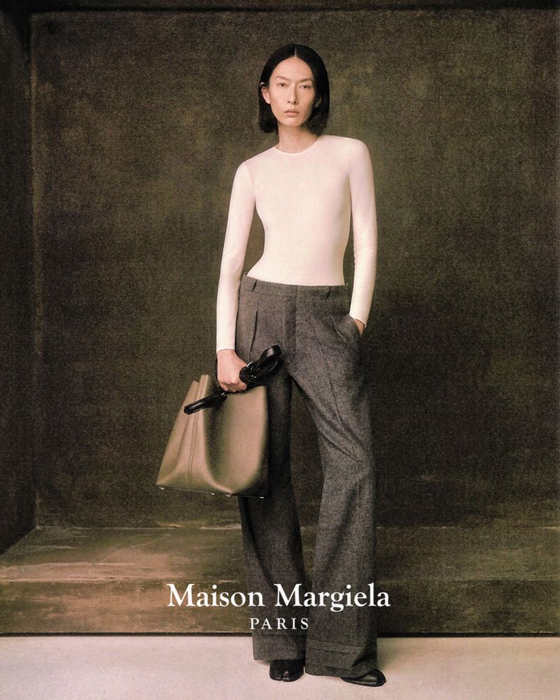 Elegant fashion shot: Asian woman poses in long-sleeve top and wool pants, holding leather bag against a textured studio backdrop.