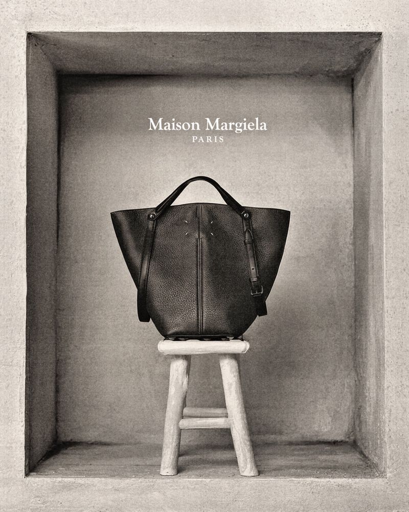 Maison Margiela black leather handbag on a pale stool against a neutral wall inside a grey niche for a studio product shot.