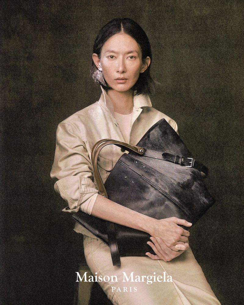 Elegant woman poses indoors with Maison Margiela handbag, wearing a light beige jacket and statement earrings against a muted backdrop.