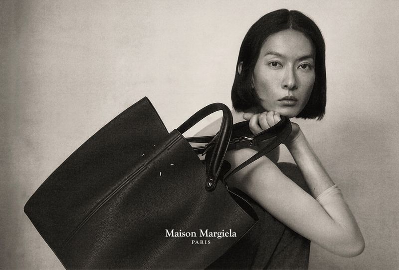 Portrait of an Asian female model posing with a black leather Maison Margiela tote bag against a neutral background in a fashion advertisement.