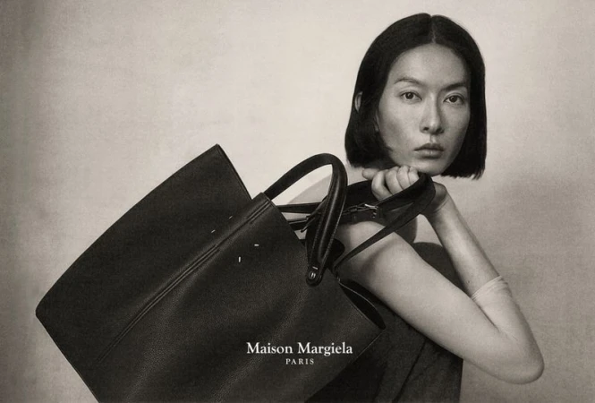 Portrait of an Asian female model posing with a black leather Maison Margiela tote bag against a neutral background in a fashion advertisement.