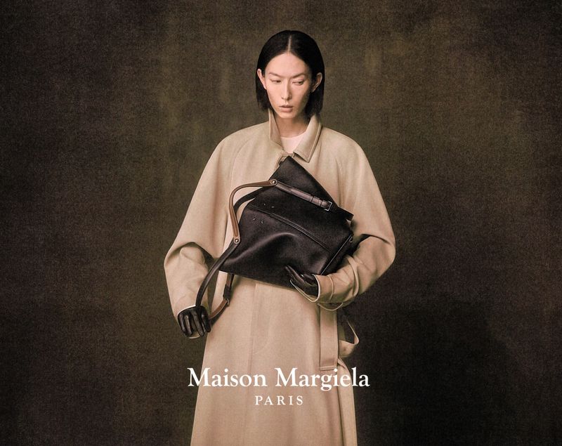 A woman with short black hair holds a black handbag while wearing a beige trench coat in a studio shot for fashion.