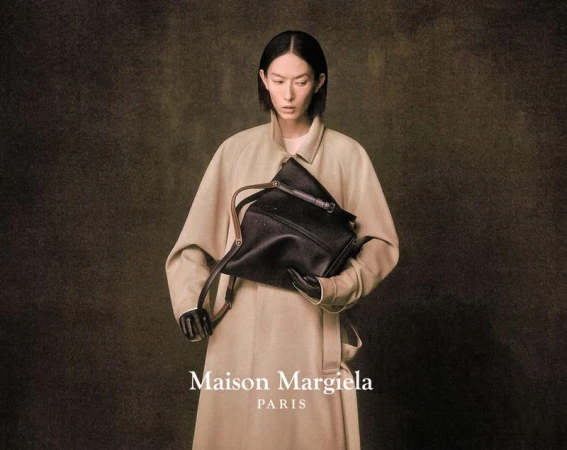 A woman with short black hair holds a black handbag while wearing a beige trench coat in a studio shot for fashion.
