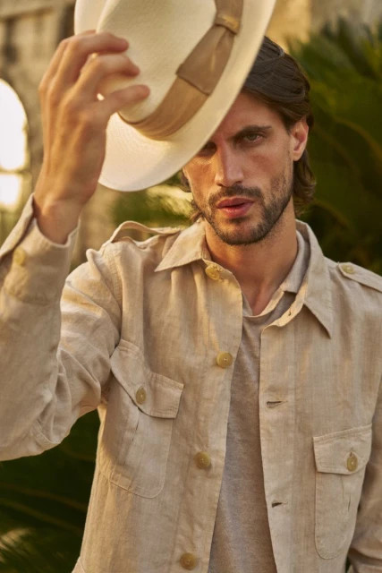 Portrait of a handsome adult male with dark hair holding a light-colored fedora hat and wearing a buttoned linen shirt outdoors.
