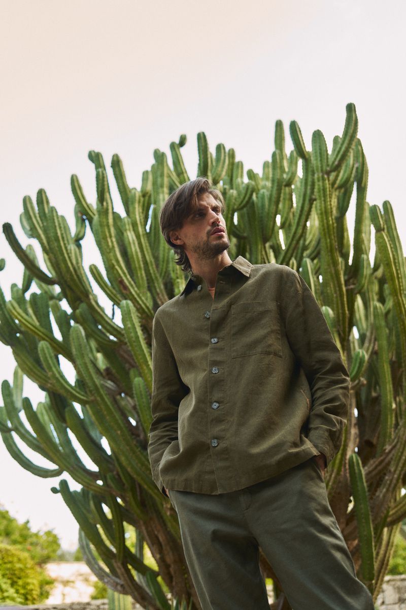 Adult man in olive shirt and trousers posing outdoors next to cactus plant, looking up on a sunny day. Fashion editorial shot.