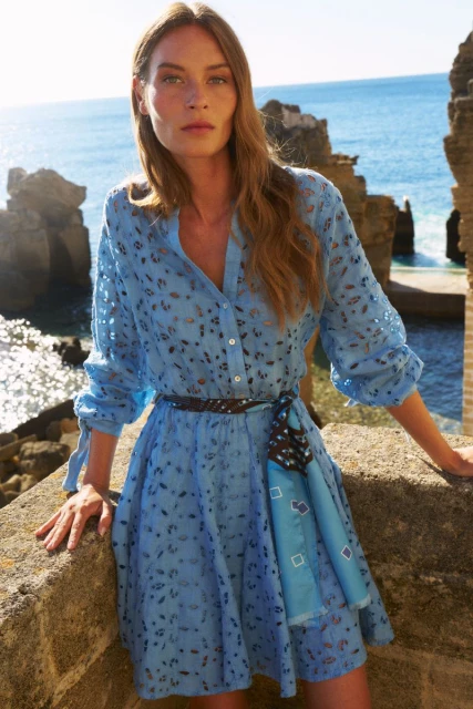 A fair-skinned woman in a light blue eyelet dress poses in front of an ocean view, complemented by rocky formations and clear skies.