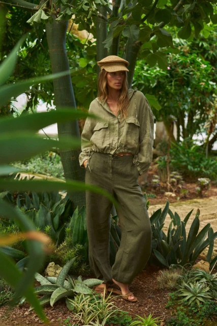 A woman in a straw cap and green linen outfit stands in a lush garden, surrounded by greenery and dappled light in a fashion editorial.