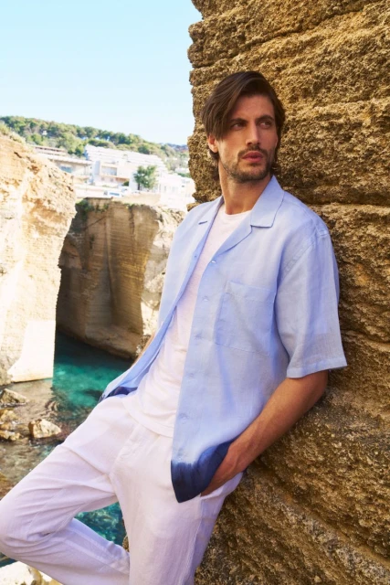 Adult man in casual coastal fashion, light blue shirt and white pants, poses against rock formation with turquoise water backdrop.