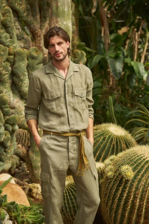 Man in casual button-down shirt and trousers poses amid a lush garden of cacti and succulents, bathed in soft, natural light, outdoor.
