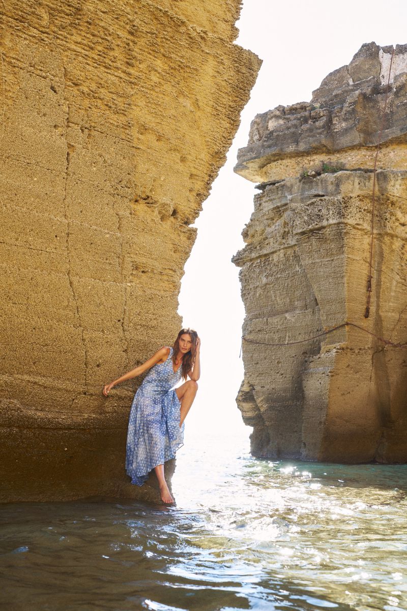 Young woman in a blue floral dress poses between large rocks in shallow sea water, bathed in sunlight, creating a serene fashion editorial.