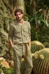 Male model wearing olive green linen shirt and matching trousers posing among various tall cacti and lush tropical plants.