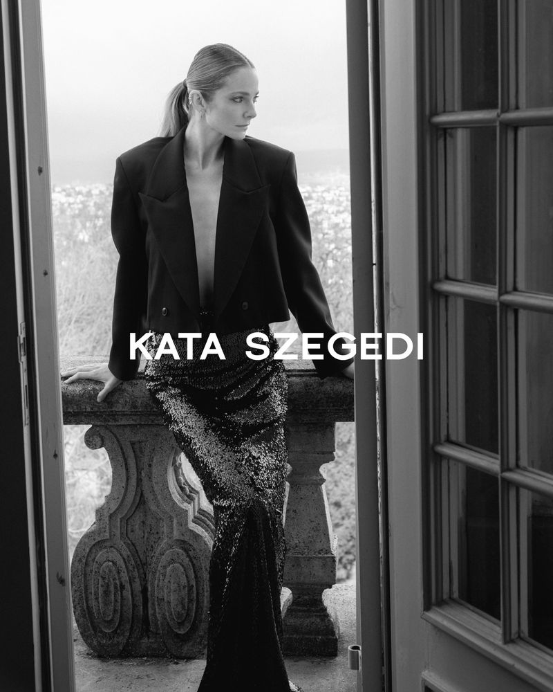 Black and white portrait of a woman in a blazer and sequined pants posing on a balcony with an urban landscape in the background.