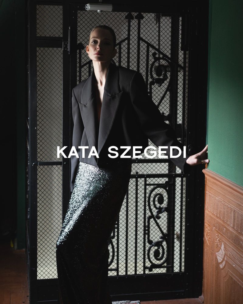 A woman in a black blazer and sparkling skirt stands in front of a dark metal door with a decorative design in an indoor setting.